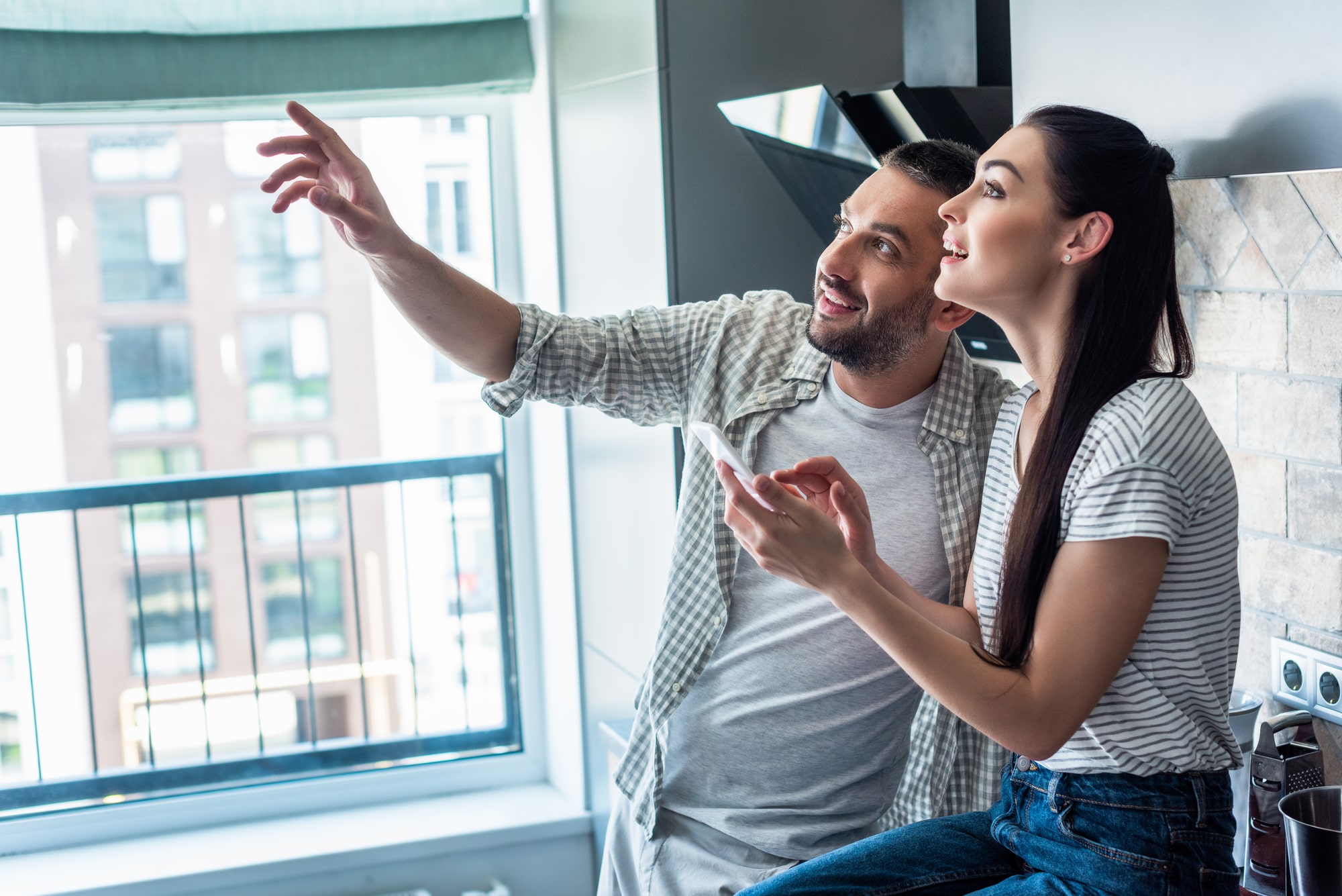 Side view of smiling couple with smartphone looking away together in kitchen, smart home